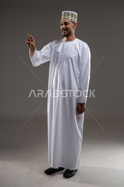 الإشارة والتوجيه الى مكان ما، بورتريه لرجل عربي خليجي عماني يرتدي الدشداشة والكمه يقف مبتسما ويشير بيده الى مكان ما، تحديد موقع  او هدف معين، حركات واشارات، خلفيه رماديه