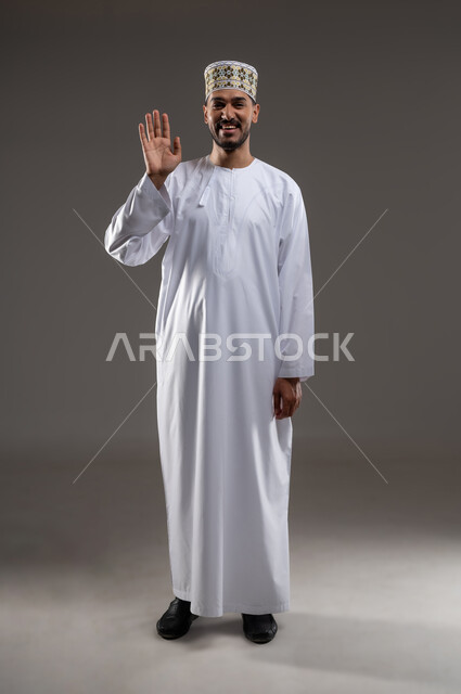 إلقاء التحية والسلام، بورتريه لرجل عربي خليجي عماني يرتدي الدشداشه والكمه يشير بيده نحو الاعلى بايماءات القاء التحيه والسلام، صورة الجسم بالطول الكامل،  خلفية رماديه
