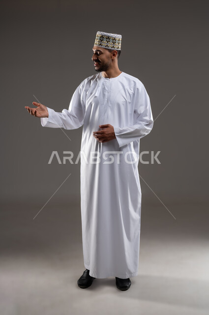 الإشارة والتوجيه الى مكان ما، بورتريه لرجل عربي خليجي عماني يرتدي الدشداشة والكمه يقف مبتسما ويشير بيده الى مكان ما، تحديد موقع  او هدف معين، حركات واشارات، خلفيه رماديه