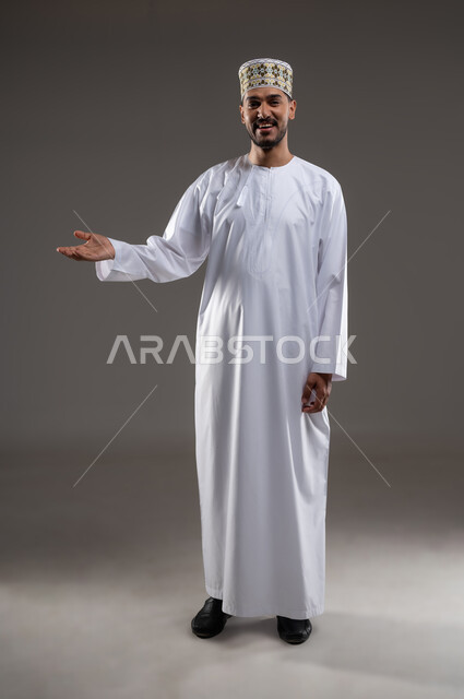 إشارات يد تدل على الترحيب الضيافه وحسن الاستقبال، بورتريه لرجل عربي خليجي عماني يرتدي الدشداشة والكمه تظهر عليه علامات الفرح والسرور يمد يده ترحيبا بضيوفه، ابتسامة لطيفة تدل على المحبة، خلفية رماديه