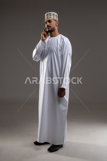 التأمل والتخطيط التمعن والتركيز، بورتريه بالطول الكامل لشاب عربي خليجي عماني يرتدي الدشداشه والكمه يشير باصبعيه نحو اذنه بايماءات وتعابير تدل على التفكير والحيرة، حركات واشارات، خلفية رمادية