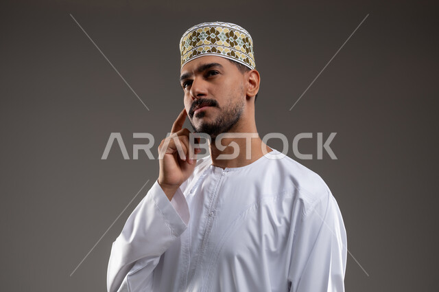 التأمل والتخطيط التمعن والتركيز، بورتريه مقرب لشاب عربي خليجي عماني يرتدي الدشداشه والكمه يشير باصبعيه نحو اذنه بايماءات وتعابير تدل على التفكير والحيرة، حركات واشارات، خلفية رمادية