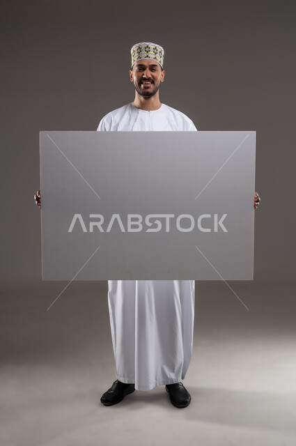 إعلانات وعروض تسويقية، استخدام لوحات توضيحية ووسائل تعليمية، بورتريه بالطول الكامل لشاب عربي خليجي عماني مبتسم يرتدي الدشداشه والكمه يحمل بيده لوح ابيض كبير (موك اب فارغ)، خلفية رماديه