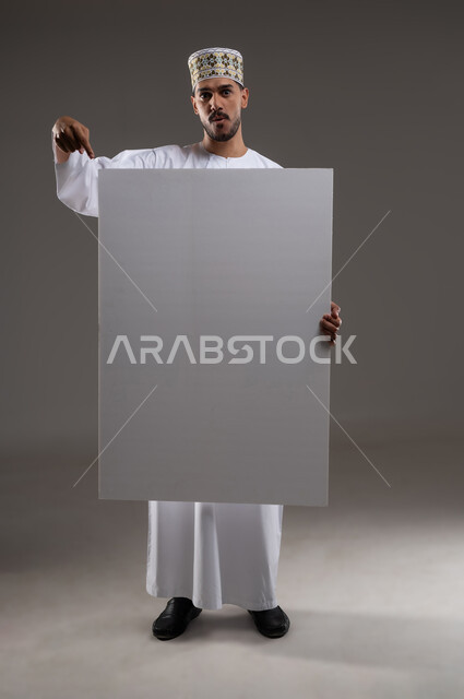 إعلانات وعروض تسويقية، استخدام لوحات توضيحية ووسائل تعليمية، بورتريه بالطول الكامل لشاب عربي خليجي عماني مبتسم يرتدي الدشداشه والكمه يحمل بيده لوح ابيض كبير (موك اب فارغ)، خلفية رماديه