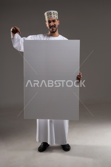 إعلانات وعروض تسويقية، استخدام لوحات توضيحية ووسائل تعليمية، بورتريه بالطول الكامل لشاب عربي خليجي عماني مبتسم يرتدي الدشداشه والكمه يحمل بيده لوح ابيض كبير (موك اب فارغ)، خلفية رماديه