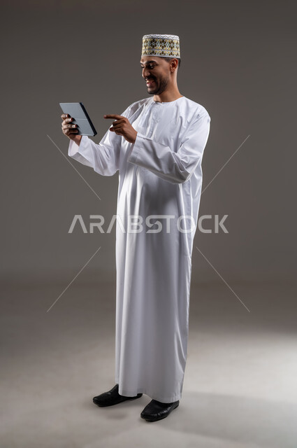 هواية المطالعة والاستكشاف وقضاء وقت الفراغ بالتعلم والثقافة، مفهوم التثقيف والبحث العلمي، بورتريه لرجل عربي خليجي عماني مبتسم يرتدي الدشداشه والكمه يحمل بيده جهاز التابلت المحمول ويقوم بالعمل عليه، خلفية رمادية