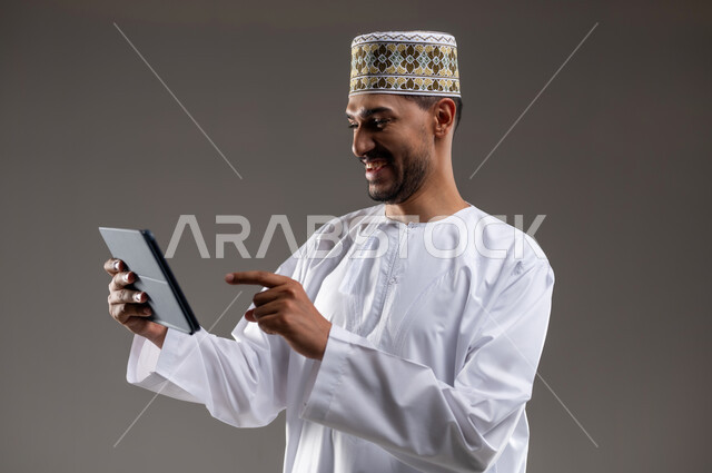 هواية المطالعة والاستكشاف وقضاء وقت الفراغ بالتعلم والثقافة، مفهوم التثقيف والبحث العلمي، بورتريه لرجل عربي خليجي عماني مبتسم يرتدي الدشداشه والكمه يحمل بيده جهاز التابلت المحمول ويقوم بالعمل عليه، خلفية رمادية