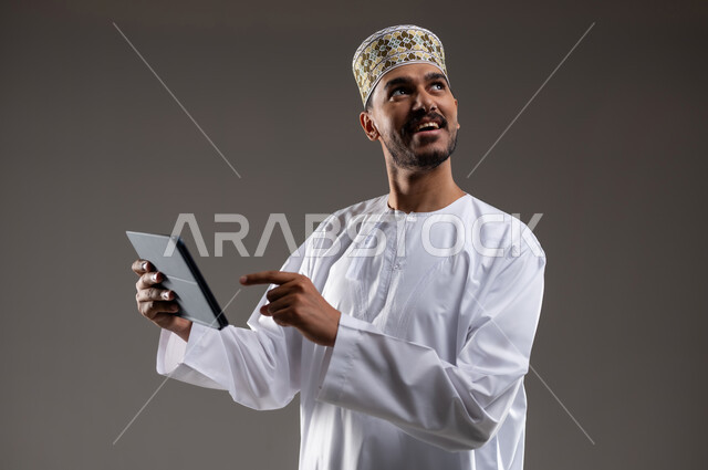 هواية المطالعة والاستكشاف وقضاء وقت الفراغ بالتعلم والثقافة، مفهوم التثقيف والبحث العلمي، بورتريه لرجل عربي خليجي عماني مبتسم يرتدي الدشداشه والكمه يحمل بيده جهاز التابلت المحمول ويقوم بالعمل عليه، خلفية رمادية
