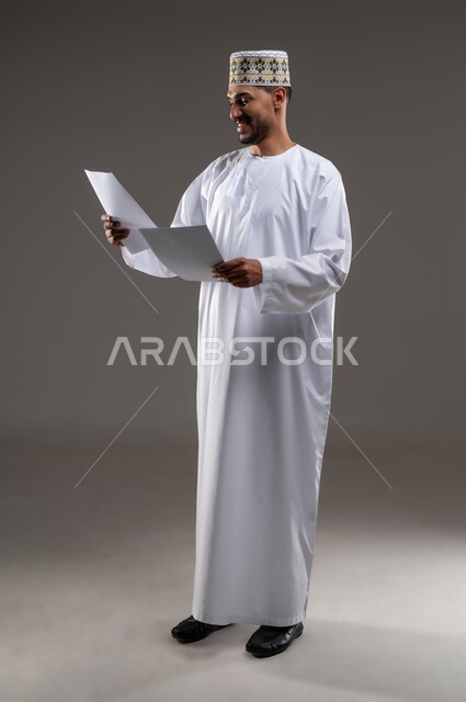 القراءه والمطالعه ودراسه المشاريع، بورتريه لرجل عربي خليجي عماني يرتدي الدشداشه والكمه يقوم بتصفح أوراقا بيضاء، قضاء أوقات الفراغ في التثقيف والتعليم، مفهوم التعليم الذاتي وممارسة الهوايات الممتعة والمفيدة في المنزل, خلفية رماديه