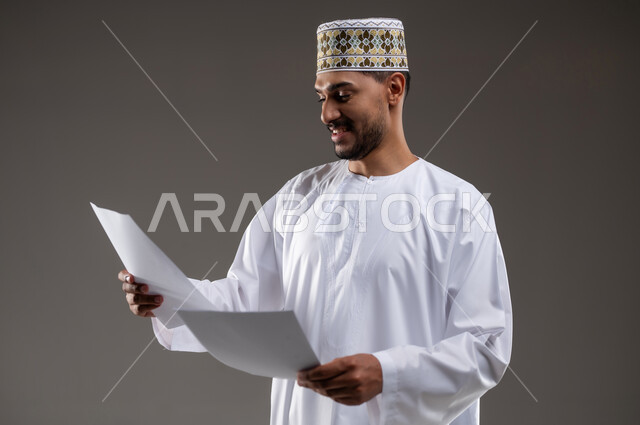 القراءه والمطالعه ودراسه المشاريع، بورتريه لرجل عربي خليجي عماني يرتدي الدشداشه والكمه يقوم بتصفح أوراقا بيضاء، قضاء أوقات الفراغ في التثقيف والتعليم، مفهوم التعليم الذاتي وممارسة الهوايات الممتعة والمفيدة في المنزل, خلفية رماديه