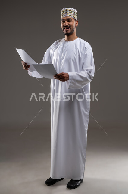 القراءه والمطالعه ودراسه المشاريع، بورتريه لرجل عربي خليجي عماني يرتدي الدشداشه والكمه يقوم بتصفح أوراقا بيضاء، قضاء أوقات الفراغ في التثقيف والتعليم، مفهوم التعليم الذاتي وممارسة الهوايات الممتعة والمفيدة في المنزل, خلفية رماديه