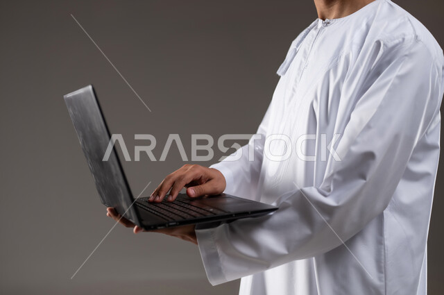 استخدام التكنولوجيا المتطورة في التسويق الرقمي الالكتروني، عرض تطبيقات وخدمات جهاز الحاسوب، بورتريه مقرب لرجل عربي خليجي عماني يرتدي الدشداشة و الكمه يحمل بيده جهاز الحاسب المحمول ويقوم بالعمل عليه، خلفية رماديه