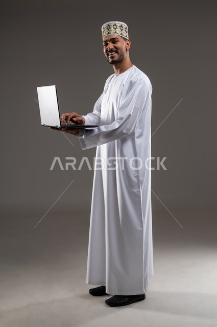 استخدام التكنولوجيا المتطورة في التسويق الرقمي الالكتروني، عرض تطبيقات وخدمات جهاز الحاسوب، بورتريه بالطول الكامل لرجل عربي خليجي عماني يرتدي الدشداشة و الكمه يحمل بيده جهاز الحاسب المحمول ويقوم بالعمل عليه، خلفية رماديه