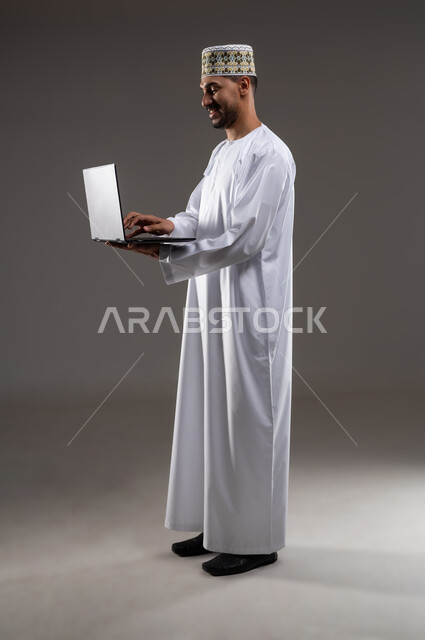 استخدام التكنولوجيا المتطورة في التسويق الرقمي الالكتروني، عرض تطبيقات وخدمات جهاز الحاسوب، بورتريه بالطول الكامل لرجل عربي خليجي عماني يرتدي الدشداشة و الكمه يحمل بيده جهاز الحاسب المحمول ويقوم بالعمل عليه، خلفية رماديه