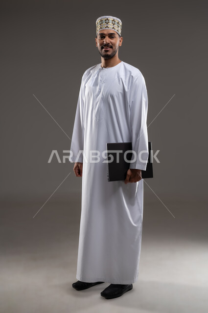 هواية المطالعة والاستكشاف وقضاء وقت الفراغ بالتعلم والثقافة، مفهوم التثقيف والبحث العلمي، بورتريه لرجل عربي خليجي عماني مبتسم يرتدي الدشداشه والكمه يحمل بيده جهاز التابلت المحمول, النظر الى الكاميرا بإيماءات الفرح والسرور، خلفية رمادية