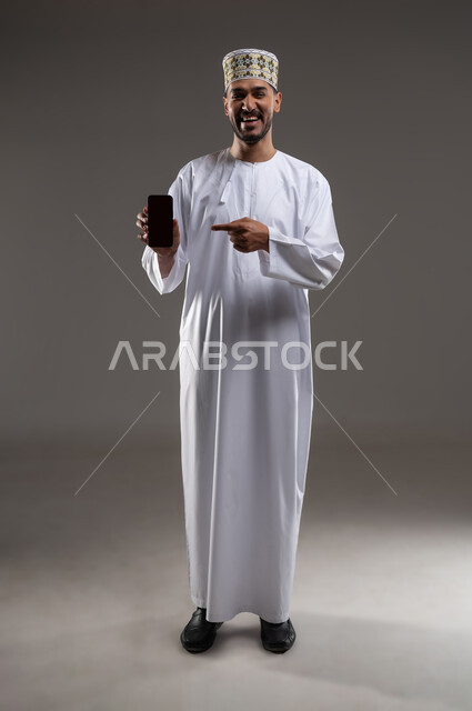 الاشارة إلى الهاتف المحمول بإيماءات السرور, بورتريه لرجل عربي خليجي عماني يرتدي الدشداشه والكمه يمسك بيده هاتفه المحمول بشاشه سوداء فارغه ويشير اليه بابتسامه, التسوق الالكتروني عبر الانترنت, استخدام التقنيات الحديثة المتطورة, خلفيه رماديه