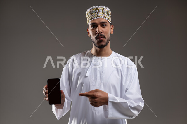 الاشارة إلى الهاتف المحمول بإيماءات السرور, بورتريه لرجل عربي خليجي عماني يرتدي الدشداشه والكمه يمسك بيده هاتفه المحمول بشاشه سوداء فارغه ويشير اليه بابتسامه, التسوق الالكتروني عبر الانترنت, استخدام التقنيات الحديثة المتطورة, خلفيه رماديه