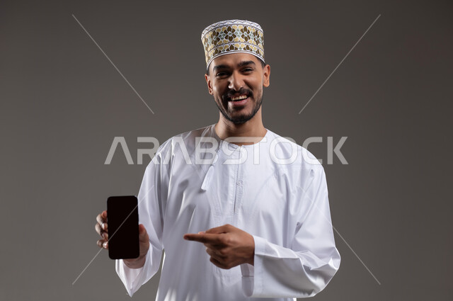 الاشارة إلى الهاتف المحمول بإيماءات السرور, بورتريه لرجل عربي خليجي عماني يرتدي الدشداشه والكمه يمسك بيده هاتفه المحمول بشاشه سوداء فارغه ويشير اليه بابتسامه, التسوق الالكتروني عبر الانترنت, استخدام التقنيات الحديثة المتطورة, خلفيه رماديه