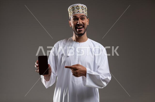 الاشارة إلى الهاتف المحمول بإيماءات السرور, بورتريه لرجل عربي خليجي عماني يرتدي الدشداشه والكمه يمسك بيده هاتفه المحمول بشاشه سوداء فارغه ويشير اليه بابتسامه, التسوق الالكتروني عبر الانترنت, استخدام التقنيات الحديثة المتطورة, خلفيه رماديه