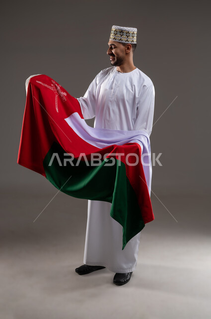 الفخر والاعتزاز بالهوية الوطنية، حب الوطن والانتماء إليه، إحياء ذكرى تأسيس الدولة 1775 م، العلم الوطني للبلاد، بورتريه  لرجل عربي خليجي عماني يرتدي الدشداشة و الكمة العمانية التقليدية يحمل بيديه العلم العماني احتفالا بذكرى اليوم الوطني 18 نوفمبر، خلفية رماديه