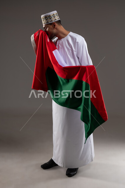 الفخر والاعتزاز بالهوية الوطنية، حب الوطن والانتماء إليه، إحياء ذكرى تأسيس الدولة 1775 م، العلم الوطني للبلاد، بورتريه  لرجل عربي خليجي عماني يرتدي الدشداشة و الكمة العمانية التقليدية يقبل العلم العماني احتفالا بذكرى اليوم الوطني العماني 18 نوفمبر، خلفية رماديه
