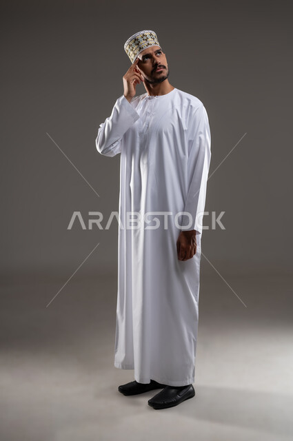 النظر الى الاعلى والتفكير بشئ ما، بورتريه لشاب عربي خليجي عماني يرتدي الدشداشه والكمه يضع يده على رأسه وينظر للاعلى بايماءات التفكير والتخطيط والتمعن في حل مشكله ما، خلفيه رماديه
