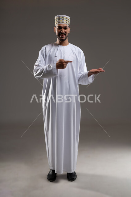 الإشارة والتوجيه الى مكان ما، بورتريه لرجل عربي خليجي عماني يرتدي الدشداشة والكمه يقف مبتسما ويشير بيده الى مكان ما، تحديد موقع  او هدف معين، حركات واشارات، خلفيه رماديه