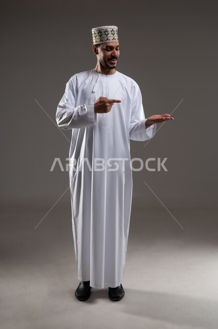 الإشارة والتوجيه الى مكان ما، بورتريه لرجل عربي خليجي عماني يرتدي الدشداشة والكمه يقف مبتسما ويشير بيده الى مكان ما، تحديد موقع  او هدف معين، حركات واشارات، خلفيه رماديه