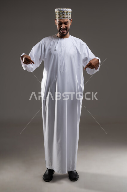 الإشارة والتوجيه الى مكان ما، بورتريه لرجل عربي خليجي عماني يرتدي الدشداشة والكمه يقف مبتسما ويشير بيده الى مكان ما، تحديد موقع  او هدف معين، حركات واشارات، خلفيه رماديه