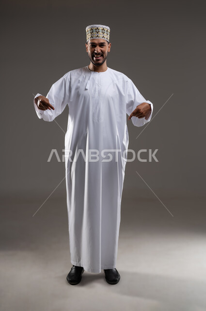 الإشارة والتوجيه الى مكان ما، بورتريه لرجل عربي خليجي عماني يرتدي الدشداشة والكمه يقف مبتسما ويشير بيده الى مكان ما، تحديد موقع  او هدف معين، حركات واشارات، خلفيه رماديه