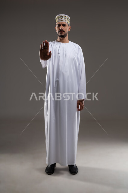 مفهوم الرفض والامتناع والانزعاج, ايماءات الحزم والجدية, طلب التوقف وعدم الاقتراب, بورتريه  لرجل عربي خليجي عماني يرتدي الدشداشة والكمه يشير بيده نحو الأعلى بتعابير النهي عن فعل امر ما, خلفية رماديه