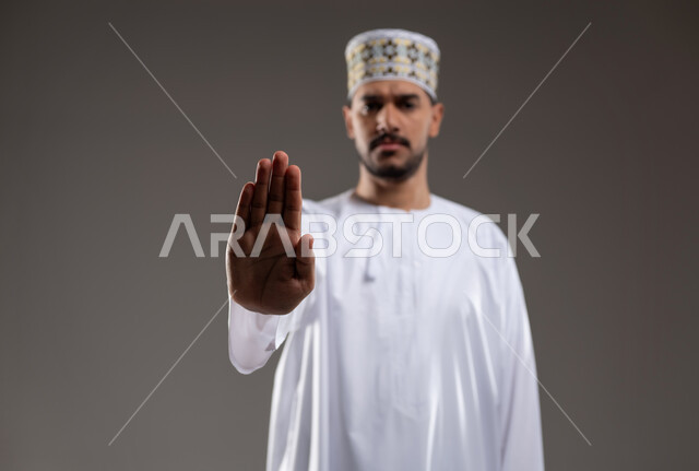 مفهوم الرفض والامتناع والانزعاج, ايماءات الحزم والجدية, طلب التوقف وعدم الاقتراب, بورتريه  لرجل عربي خليجي عماني يرتدي الدشداشة والكمه يشير بيده نحو الأعلى بتعابير النهي عن فعل امر ما, خلفية رماديه