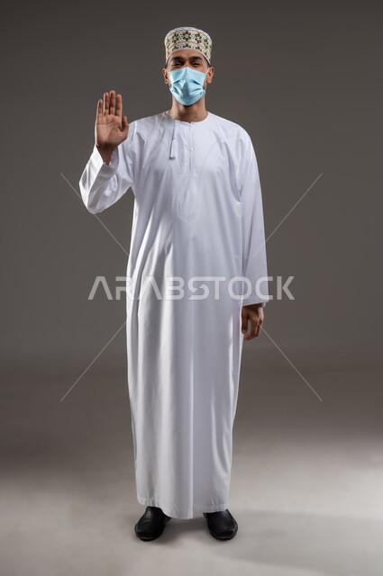 Wearing a mask to prevent the Corona virus Covid-19, maintaining distances and avoiding superficial contact, portrait of a young Arab Gulf Omani man wearing a dishdasha, a kimono and a medical mask, pointing his palm upwards with gestures of avoiding shaking hands, not getting close and adhering to preventive measures, gray background