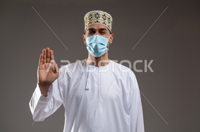 ارتداء الكمامة للوقاية من فايروس كورونا كوفيد-19، الحفاظ على المسافات وتجنب الملامسة السطحية، بورتريه  لشاب عربي خليجي عماني يرتدي الدشداشة والكمه والكمامه الطبيه يشير بكف يده نحو الاعلى بايماءات تجنب المصافحة وعدم الاقتراب و الالتزام بالإجراءات الوقائية، خلفية رماديه