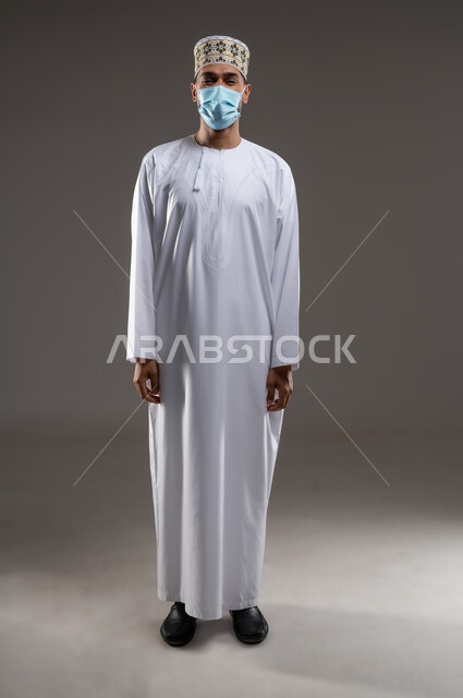 ارتداء الكمامة للوقاية من فايروس كورونا كوفيد-19، الحفاظ على المسافات وتجنب الملامسة السطحية، بورتريه  لشاب عربي خليجي عماني يرتدي الدشداشة والكمه والكمامه الطبيه يقف مرتديا كمامته الطبيه بايماءات الالتزام بالإجراءات الوقائية، خلفية رماديه