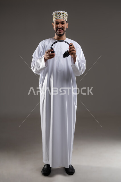 Wearing headphones and interacting with the music, gestures and expressions of integration with the song, portrait of an Arab Gulf Omani man wearing a dishdasha and a kummah wearing headphones to listen to music, gestures indicating listening and interacting with the music, gray background