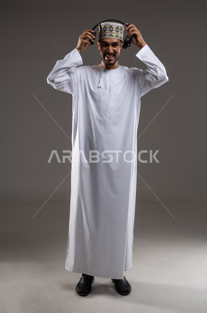 Wearing headphones and interacting with the music, gestures and expressions of integration with the song, portrait of an Arab Gulf Omani man wearing a dishdasha and a kummah wearing headphones to listen to music, gestures indicating listening and interacting with the music, gray background