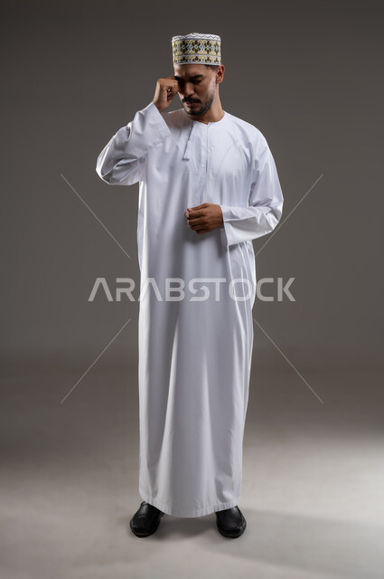 الشعور بالقهر والخذلان الحزن والبكاء، بورتريه لشاب عربي خليجي عماني يرتدي الدشداشه والكمه يقوم بفرك عينيه بيديه بايماءات الحزن والخساره، خلفيه رماديه