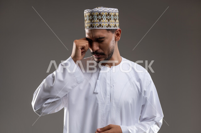 الشعور بالقهر والخذلان الحزن والبكاء، بورتريه لشاب عربي خليجي عماني يرتدي الدشداشه والكمه يقوم بفرك عينيه بيديه بايماءات الحزن والخساره، خلفيه رماديه