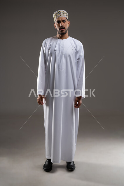 الإندهاش و المفاجأة من شيء ما، بورتريه لرجل عربي خليجي عماني يرتدي الدشداشه والكمه يقف باستقامه بايماءات الدهشه والاستغراب، خلفية رماديه