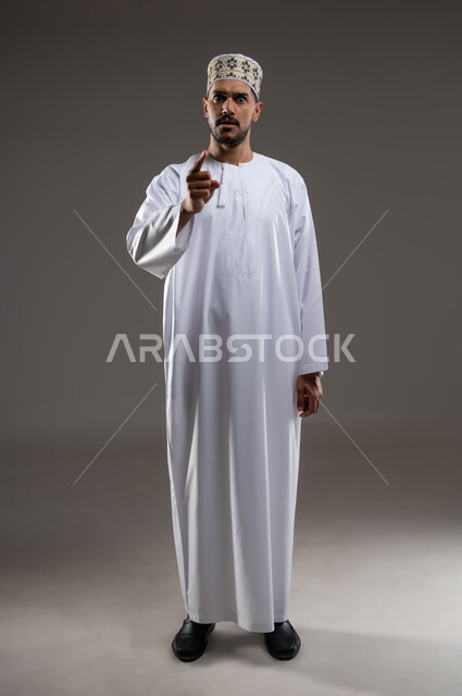 الإشارة والتحذير من شئ ما، بورتريه لرجل عربي خليجي عماني يرتدي الدشداشة والكمه يقف ويشير باصبع السبابه بأيماءات الجديه والتهديد والغضب، التوعد بتنفيذ أمر ما، حركات واشارات، خلفيه رماديه