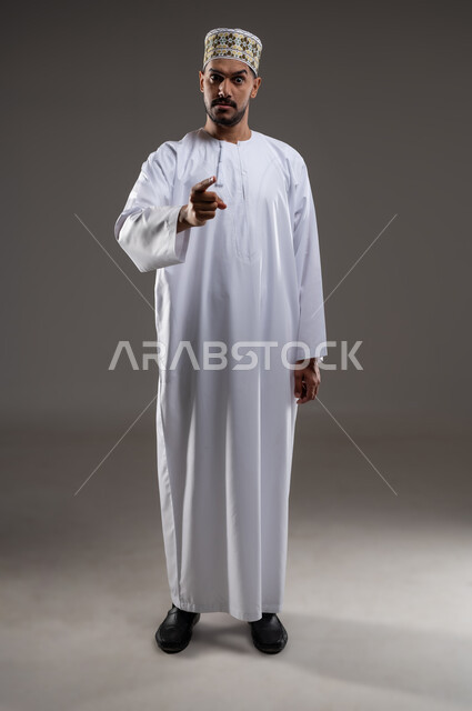 الإشارة والتحذير من شئ ما، بورتريه لرجل عربي خليجي عماني يرتدي الدشداشة والكمه يقف ويشير باصبع السبابه بأيماءات الجديه والتهديد والغضب، التوعد بتنفيذ أمر ما، حركات واشارات، خلفيه رماديه
