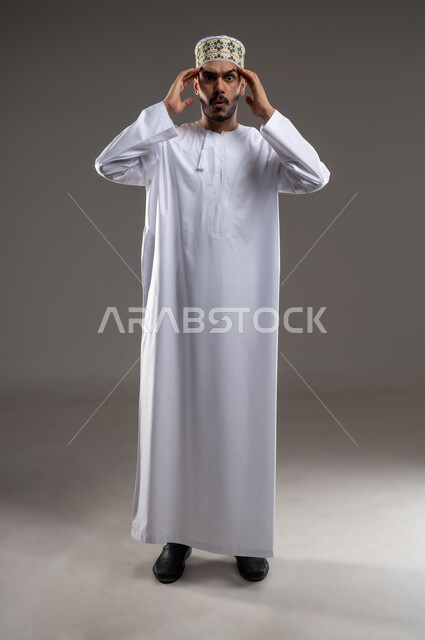 مفهوم الحاجة الى الهدوء، بورتريه لشاب عربي خليجي عماني يرتدي الدشداشة و الكمه يضع يديه على رأسه بتعابير التفكير والحاجه الى الراحه، حل المشكلات واتخاذ القرارات, إيماءات التمعن والتركيز والتخطيط للمستقبل، خلفيه رماديه