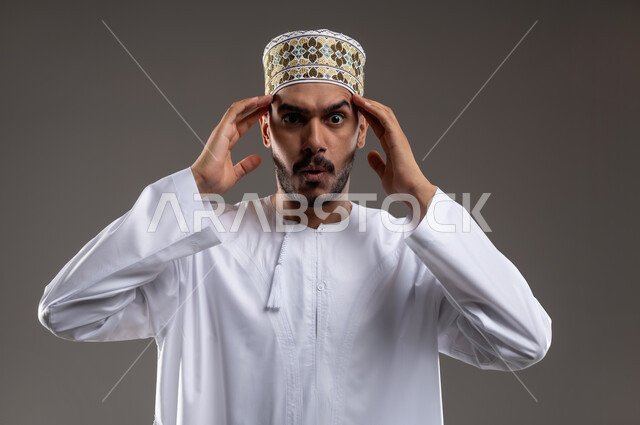 مفهوم الحاجة الى الهدوء، بورتريه لشاب عربي خليجي عماني يرتدي الدشداشة و الكمه يضع يديه على رأسه بتعابير التفكير والحاجه الى الراحه، حل المشكلات واتخاذ القرارات, إيماءات التمعن والتركيز والتخطيط للمستقبل، خلفيه رماديه