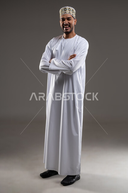 مفهوم الرجولة والثقة بالنفس، التقاط الصور الشخصية للذكريات، بورتريه مقرب لشاب عربي خليجي عماني مبتسم يرتدي الدشداشة و الكمه ينظر الى الكاميرا بإيماءات السرور والسعادة مكتوف اليدين، الاهتمام بالأناقة والمظهر الخارجي، خلفية رماديه