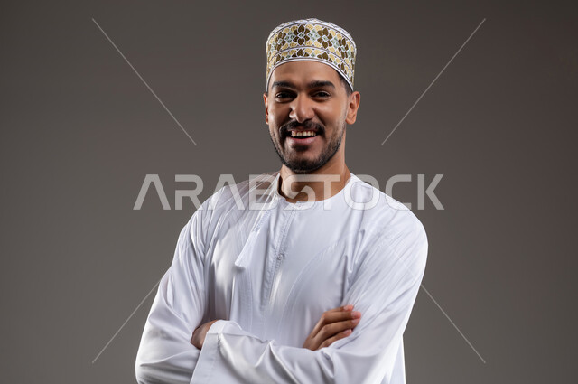 مفهوم الرجولة والثقة بالنفس، التقاط الصور الشخصية للذكريات، بورتريه مقرب لشاب عربي خليجي عماني مبتسم يرتدي الدشداشة و الكمه ينظر الى الكاميرا بإيماءات السرور والسعادة مكتوف اليدين، الاهتمام بالأناقة والمظهر الخارجي، خلفية رماديه