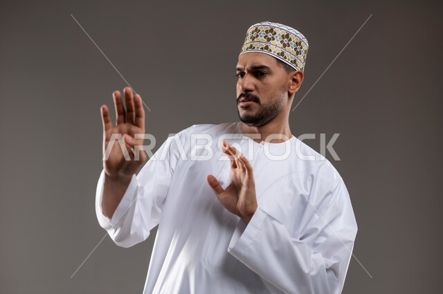 تعابير الذعر والخوف من شيء ما، مد اليدين للأمام محاولةً للدفاع عن النفس، بورتريه لرجل عربي عماني يرتدي الدشداشة والكمه تظهر عليه علامات الرعب، ايماءات تدل على طلب النجدة والانقاذ، خلفية رماديه