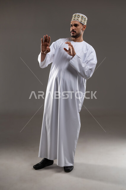 تعابير الذعر والخوف من شيء ما، مد اليدين للأمام محاولةً للدفاع عن النفس، بورتريه لرجل عربي عماني يرتدي الدشداشة والكمه تظهر عليه علامات الرعب، ايماءات تدل على طلب النجدة والانقاذ، خلفية رماديه