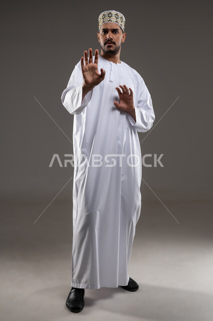 تعابير الذعر والخوف من شيء ما، مد اليدين للأمام محاولةً للدفاع عن النفس، بورتريه لرجل عربي عماني يرتدي الدشداشة والكمه تظهر عليه علامات الرعب، ايماءات تدل على طلب النجدة والانقاذ، خلفية رماديه