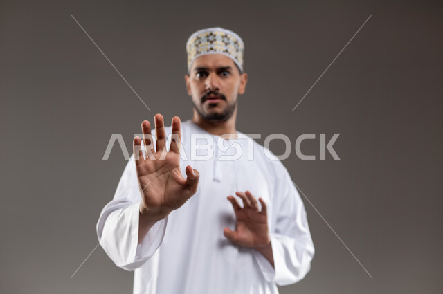 تعابير الذعر والخوف من شيء ما، مد اليدين للأمام محاولةً للدفاع عن النفس، بورتريه لرجل عربي عماني يرتدي الدشداشة والكمه تظهر عليه علامات الرعب، ايماءات تدل على طلب النجدة والانقاذ، خلفية رماديه
